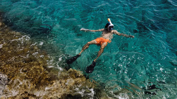 A man in orange swim shorts and snorkeling gear bobs in clear waters