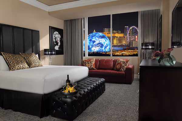 A hotel room with a large bed, a red sofa, a black ottoman, and a view of a cityscape with a Ferris wheel.