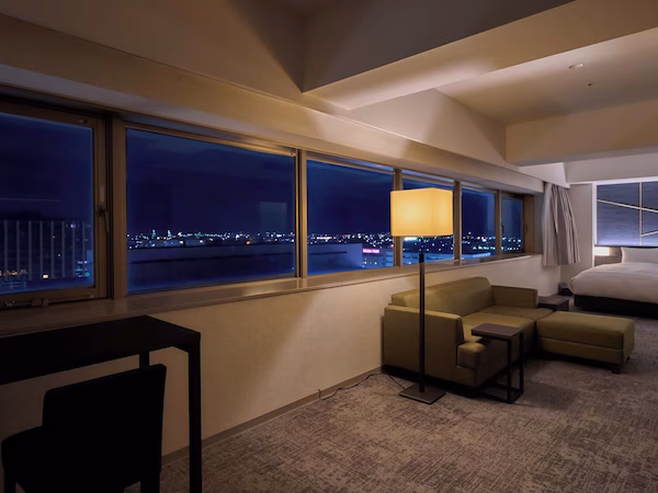 A hotel room with a large window offering a city view at night, a sofa, a desk, and a bed.