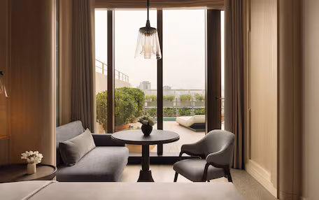 A modern hotel room with a large window, a sofa, two chairs, a small round table, and a hanging light fixture.