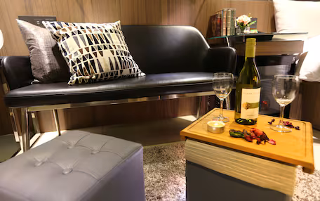 A modern living room with a black leather sofa, a white ottoman, and a wooden coffee table with a bottle of wine, glasses, and a candle.