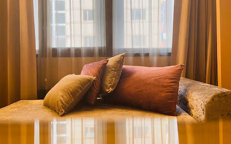 A sofa with cushions in a room with sheer curtains and a view of buildings outside.